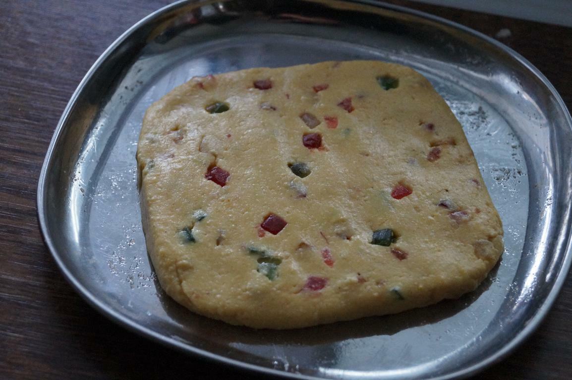 Burfi mixture spread and set in a tray with tutti frutti pieces for cutting.