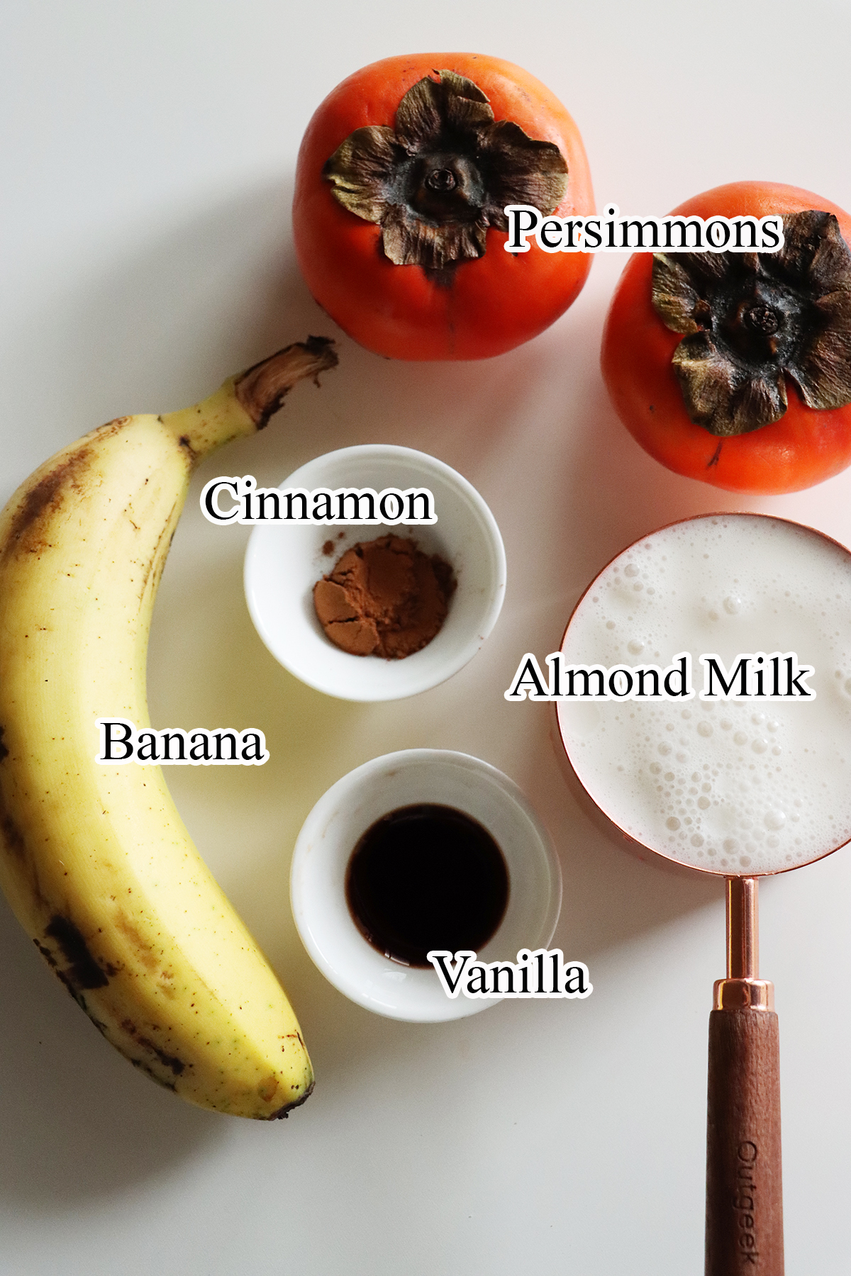 Banana, cinnamon ,almond milk, persimmons and vanilla placed on a white surface to prepare vegan persimmon smoothie