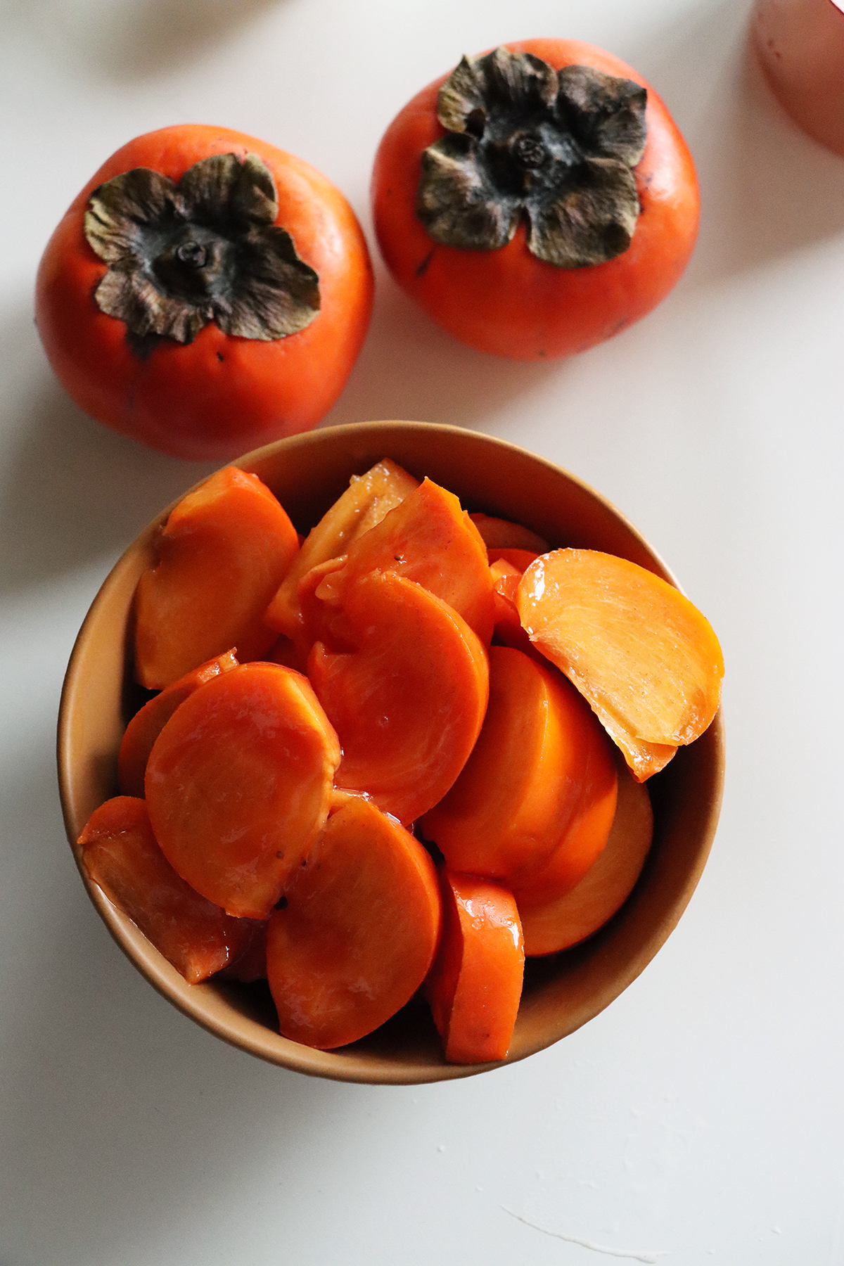 A bowl of sliced persimmons on a white table along with two whole persimmons.