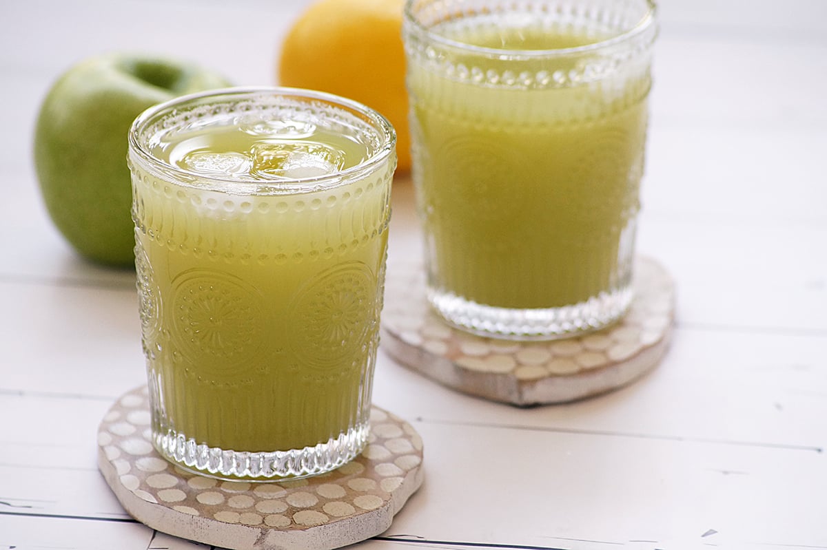 Two glasses of green apple juice placed on wooden coasters on a table