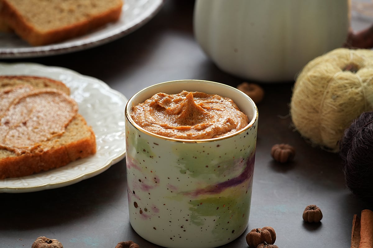 A slice of bread with pumpkin pie spice butter on a white plate, a cup of pumpkin spiced butter, surrounded by cinnamon sticks and fall decorations.
