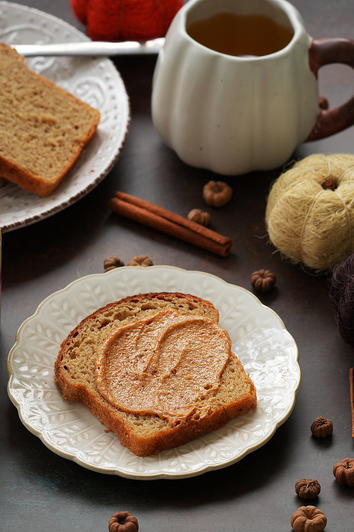 A slice of bread with pumpkin pie spice butter on a white plate, served with a cup of tea surrounded by cinnamon sticks and fall decorations.