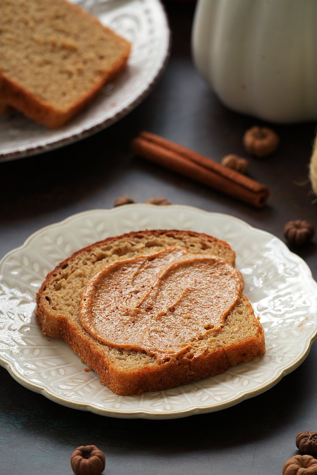 A slice of bread with pumpkin pie spice butter on a white plate, surrounded by cinnamon sticks and fall decorations.