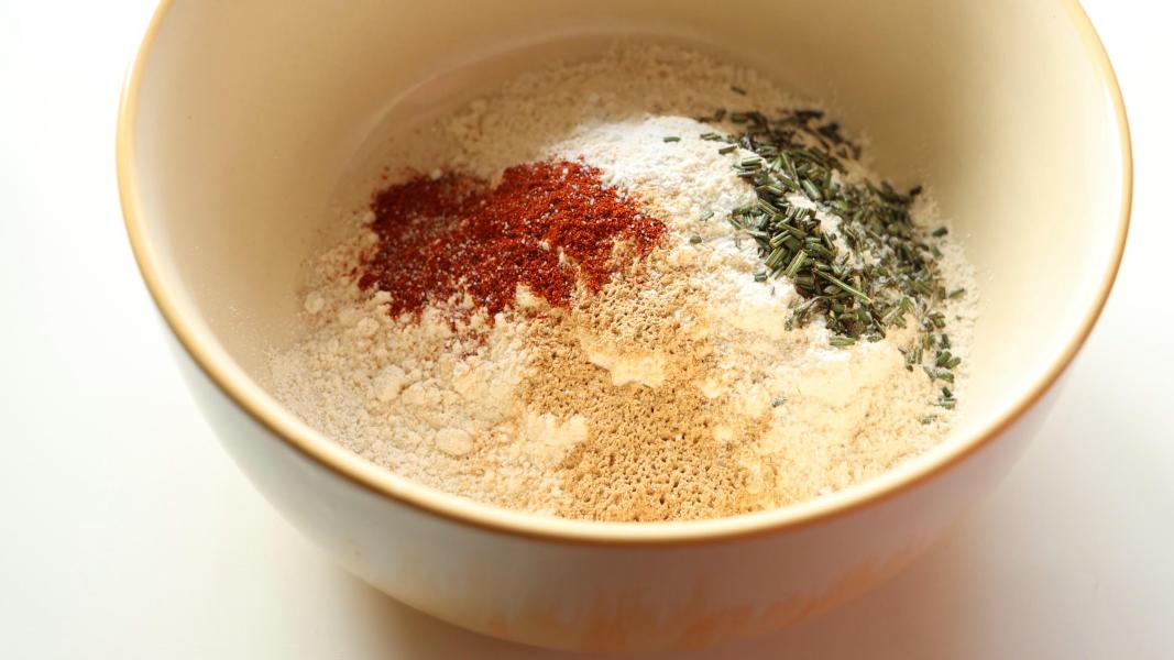 Flour, baking powder, spices, salt, and rosemary are added to a yellow ceramic mixing bowl.