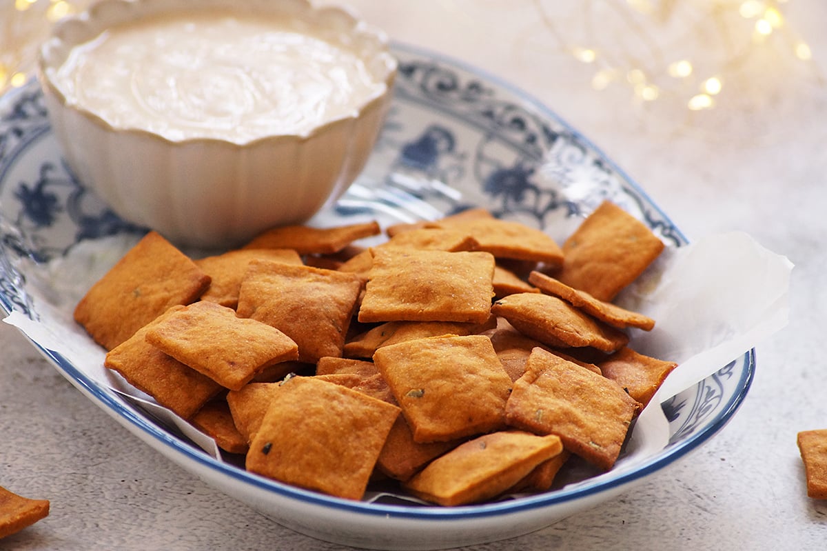 A close-up of homemade sweet potato crackers served on a blue and white plate with a creamy yogurt dip on the side.