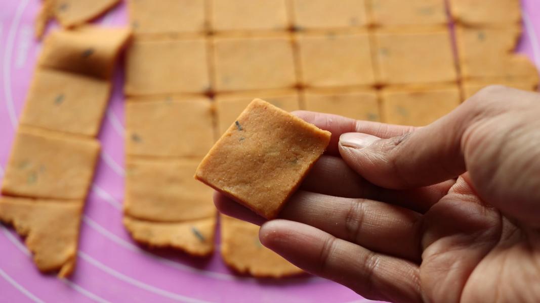 A hand holding a square piece of sweet potato cracker dough, with more dough squares on a mat in the background.