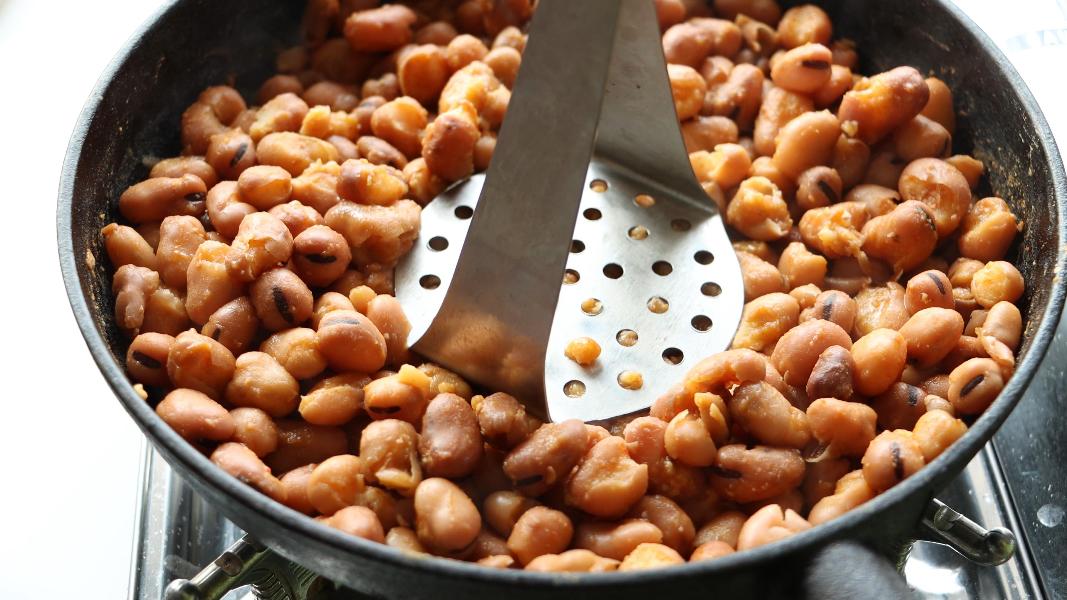 Simmered fava beans being mashed with a potato masher for making Egyptian fava beans stew