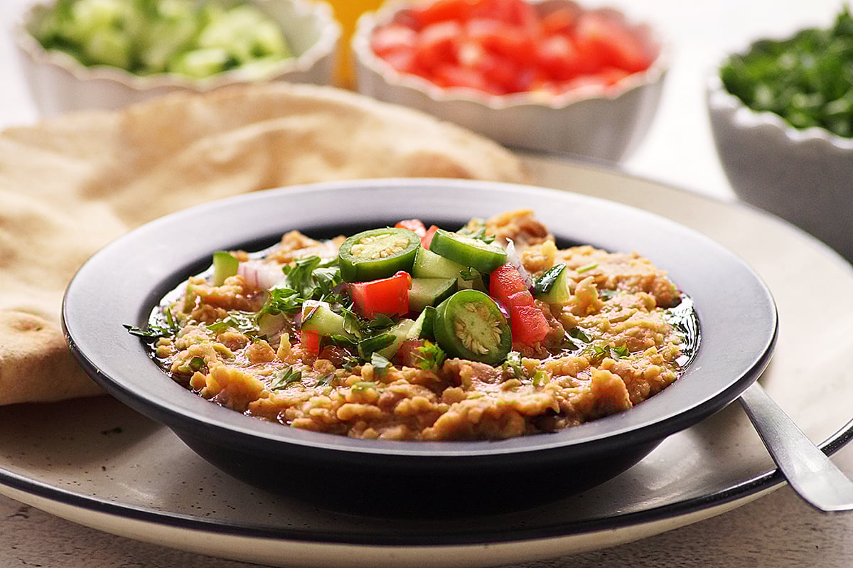 Authentic ful medames served in a bowl topped with cucumber, tomato, onion, jalapeno and olive oil with pita bread on the side.