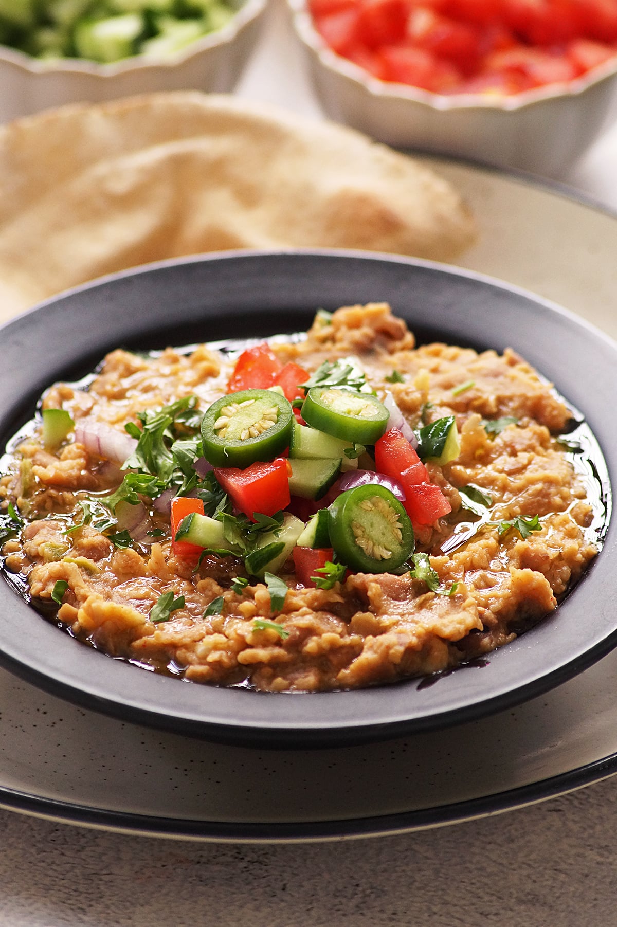 Egyptian ful medames served in a black shallow bowl, topped with chopped onion, tomato, cucumber, parsley and jalapeno. Served alongside pita bread